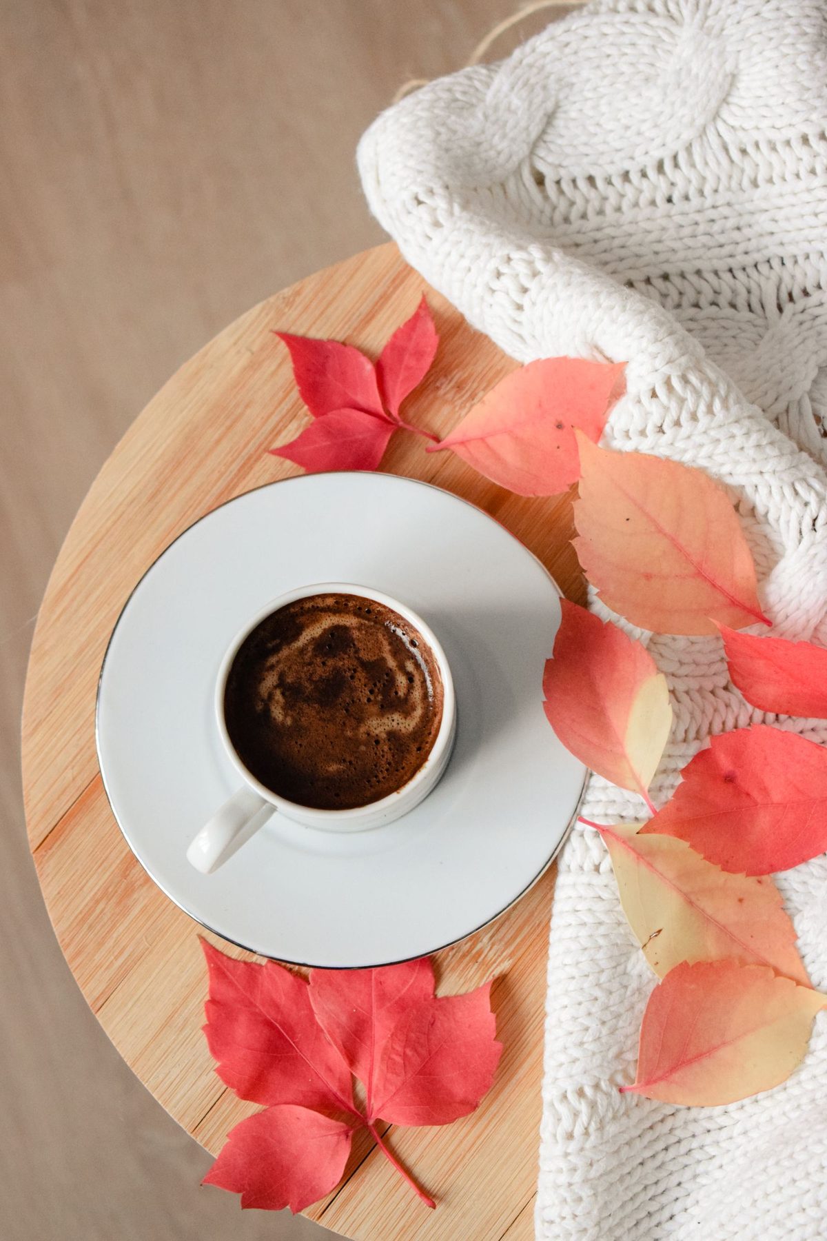 A cup of coffee on a wooden board surrounded by red autumn leaves and a knitted blanket