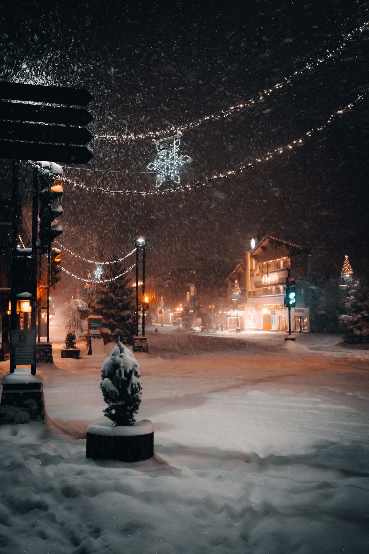A snowy town street decorated with Christmas lights as snow falls at night